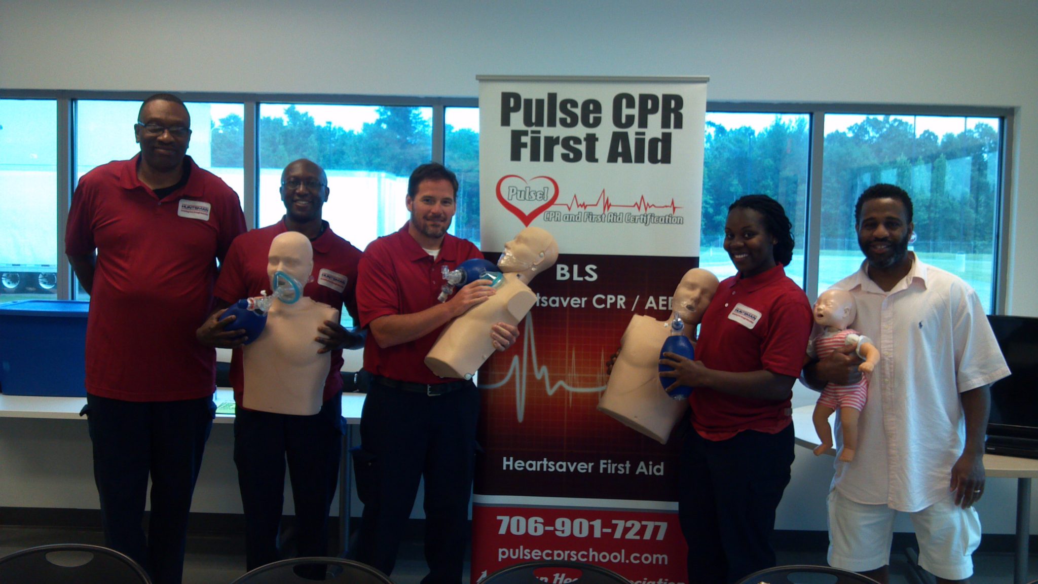 A group of men standing together, smiling while posing with a first aid training dummy in a classroom setting.