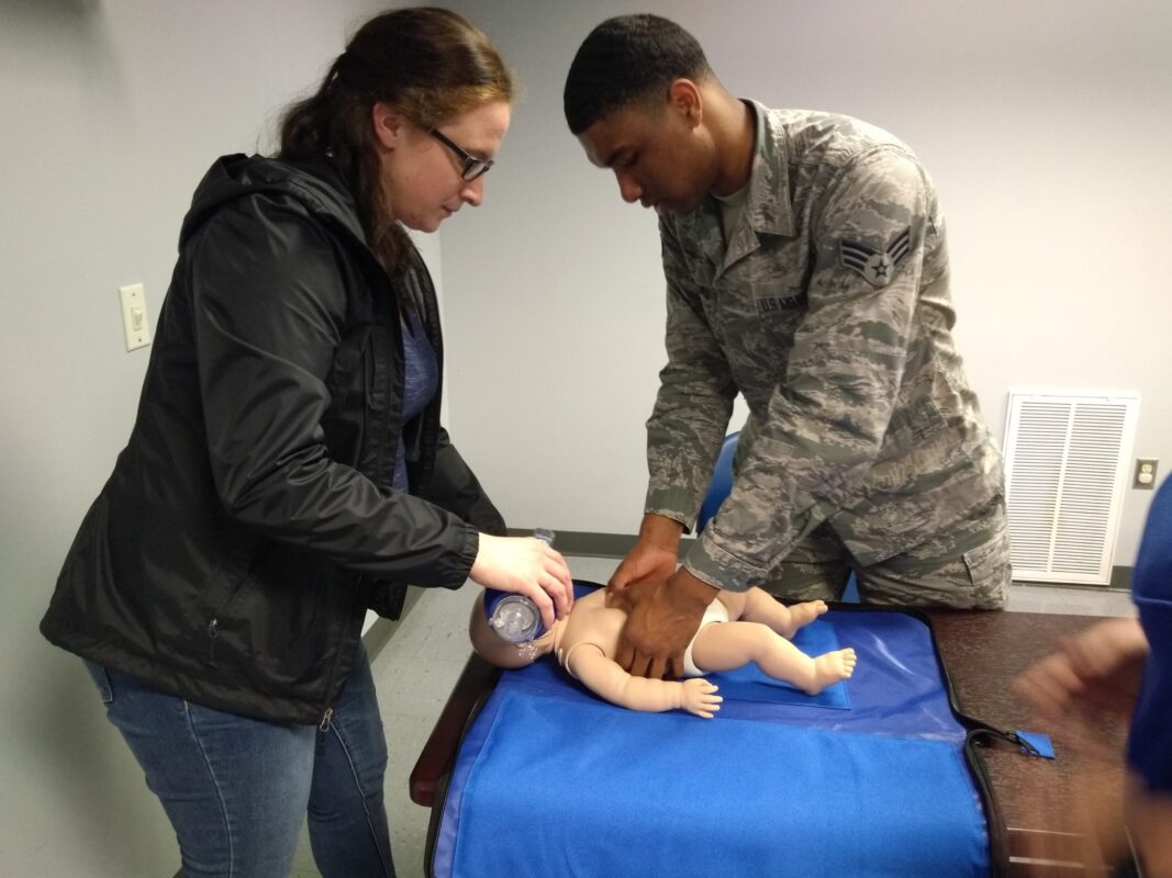 A man and woman in uniform assist a baby during CPR training at Fort Gordon for workplace safety.